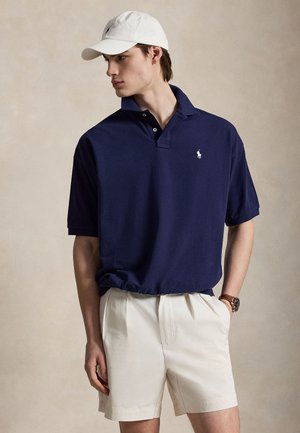 Mand iført navy polo, hvide shorts, hvid baseballkasket og armbåndsur, stående med hænderne i lommerne og ser til venstre.