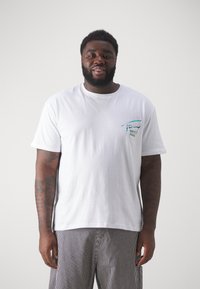 Camiseta blanca de algodón con corte holgado, que presenta un pequeño logotipo colorido en el lado izquierdo del pecho. Combinada con pantalones cortos a rayas.