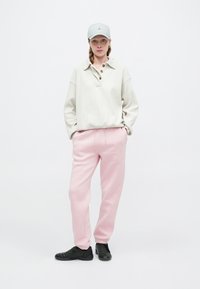 Pulover oversized gri deschis din tricot cu model waffle, cu trei butoane, asortat cu pantaloni de trening roz și pantofi sport negri; prezintă o șapcă bleu deschis.