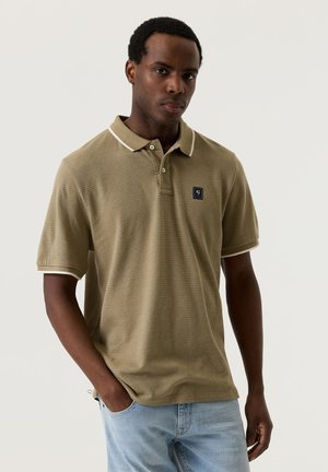 Jonge man draagt een olijfgroene, gestructureerde poloshirt met witte bies en lichtblauwe spijkerbroek, staand met één hand in zijn zak.