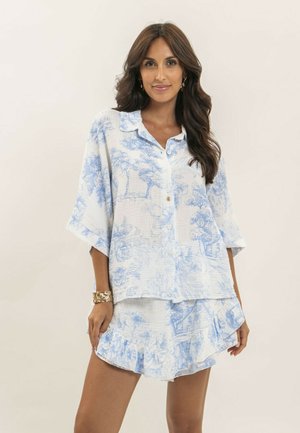 Femme portant un chemisier blanc ample avec des imprimés bleus pittoresques et un short assorti à volants, debout devant un fond clair uni.