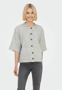 Femme souriante avec des cheveux blonds courts, portant un cardigan boutonné gris clair et un pantalon gris foncé, se tenant devant un fond uni.