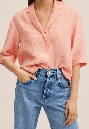 Femme portant une chemise ample à manches courtes texturée couleur pêche rentrée dans un jean en denim bleu taille haute, main dans la poche.