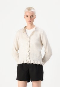 Cardigan bej cu margine ondulată, având un închidere cu butoane în față și mâneci lungi, realizat dintr-un material moale și texturat. Purtați deasupra unei bluze albe.