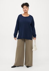 Μπλέ υπερμεγέθη μπλούζα με μακριά μανίκια και στρογγυλή λαιμόκοψη, συνδυασμένη με ανοιχτόχρωμες μπεζ παντελόνες και μία μικρή λευκή τσάντα χεριού. Κομψή, χαλαρή εφαρμογή.