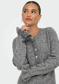Femme aux cheveux tirés en arrière, portant des boucles d'oreilles en argent et un cardigan tricoté gris à boutons, la main près du menton, regardant vers la droite.