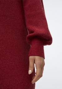 Rød strikket sweater med ribbet tekstur og lange ærmer, som har en afslappet pasform og manchetter i kanten. Nærbillede af en hånd, der hviler mod stoffet.