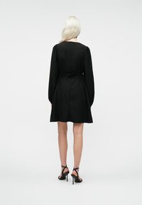 Γυναίκα με μακριά ξανθά μαλλιά, φορώντας ένα μαύρο υφασμάτινο φόρεμα μέχρι το γόνατο με μακριά μανίκια και μαύρες ψηλοτάκουνες σάντες, στέκεται με την πλάτη γυρισμένη.