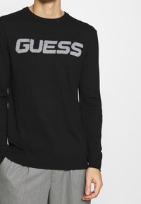 Schwarzer Strickpullover mit einem kontrastierenden grauen "GUESS"-Logo auf der Brust, mit gerippten Bündchen und Saum.
