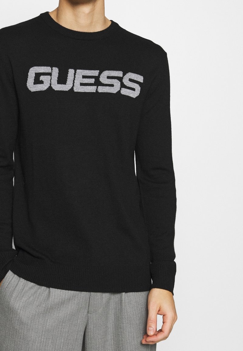Schwarzer Strickpullover mit einem kontrastierenden grauen "GUESS"-Logo auf der Brust, mit gerippten Bündchen und Saum.