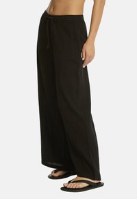 Mujer con pantalones negros holgados con cordón y chanclas negras, de pie frente a un fondo claro y liso.