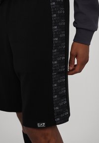 Pantaloni sportivi neri con una texture liscia, caratterizzati da un pannello laterale con testo ripetuto in grigio scuro. Include un'etichetta con logo sul fondo.