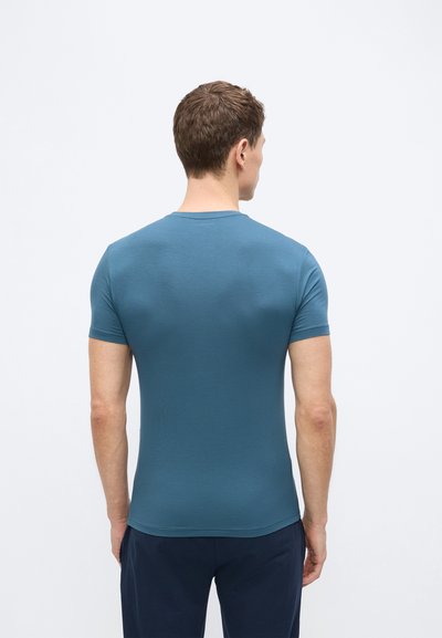 Άνδρας με την πλάτη γυρισμένη, φορώντας εφαρμοστό μπλε κοντομάνικο πουκάμισο και σκουρόχρωμο παντελόνι, μπροστά σε απλό φωτεινό φόντο.