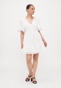 Abito bianco corto con scollatura a V, maniche rifinite in pizzo e orlo a occhiello. Tessuto leggero con una vestibilità comoda e bottoni decorativi.