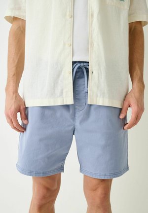 Mann trägt hellblaue Shorts mit Tunnelzug, ein weißes, geripptes Tanktop und ein cremefarbenes kurzärmliges Hemd, die Hände seitlich am Körper.