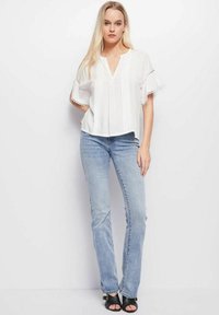 Blusa bianca a maniche corte con pieghe e bordino in pizzo, abbinata a jeans azzurri a campana e sandali neri, su uno sfondo neutro.