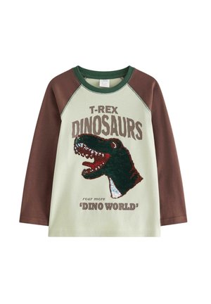 Ung dreng, der balancerer med udstrakte arme iført dinosaur t-shirt, grønne bukser og grønne og hvide sneakers på ensfarvet baggrund.