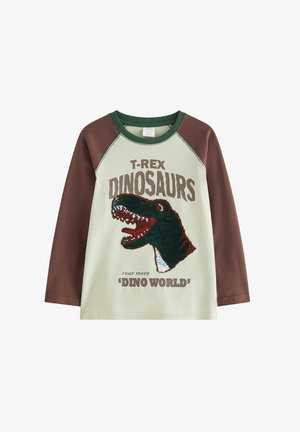 Ung dreng, der balancerer med udstrakte arme iført dinosaur t-shirt, grønne bukser og grønne og hvide sneakers på ensfarvet baggrund.