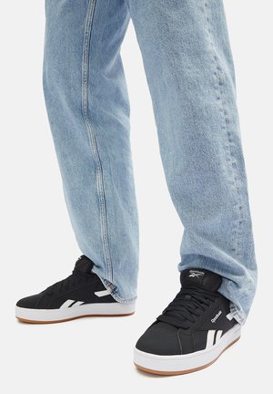 Zapatillas negras con detalles blancos y una suela de goma, con un diseño de caña alta, cordones y detalle del logo. Llevadas con jeans de color azul claro.