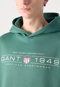 Felpa verde realizzata in tessuto morbido, con una grafica frontale che presenta "GANT 1949" e un'emblema a forma di scudo. Include un cappuccio con cordino.
