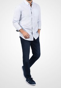 Camisa abotonada de color azul claro con mangas largas, combinada con jeans de denim oscuro y zapatillas azules oscuras, con un diseño sencillo y detalle de bolsillo.