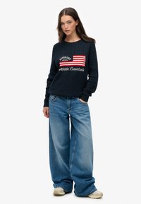 Marineblauwe kabelgebreide trui met een rood en witte gestreepte grafiek en de tekst "Athletic Essentials", gecombineerd met lichtblauwe wijde spijkerbroeken.