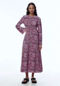 Langärmliges pinkes Kleid mit schwarzem Leopardenmuster, mit einer taillierten Taille und seitlichem Schlitz. Getragen mit klobigen schwarzen Sandalen.