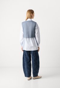 Gilet trapuntato azzurro chiaro sopra una camicia bianca, abbinato a pantaloni larghi scuri con motivo floreale e scarpe beige senza lacci. Texture morbide evidenti.