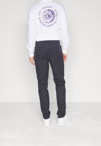 Bílé tričko s dlouhým rukávem s fialovým květinovým logem na zádech, v kombinaci s tmavě šedými úzkými kalhotami a bílými teniskami. Pohled zezadu.