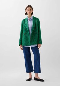 Blazer verde doppiopetto con un motivo a strisce di tigre testurizzate, abbinato a una camicia azzurro chiaro e jeans cropped blu scuro.