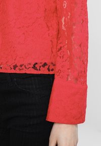 Czerwona koronkowa bluzka z długimi rękawami, zdobiona kwiatowymi wzorami i falowanym dołem. Połączona z czarnymi spodniami w paski oraz szerokim mankietem.