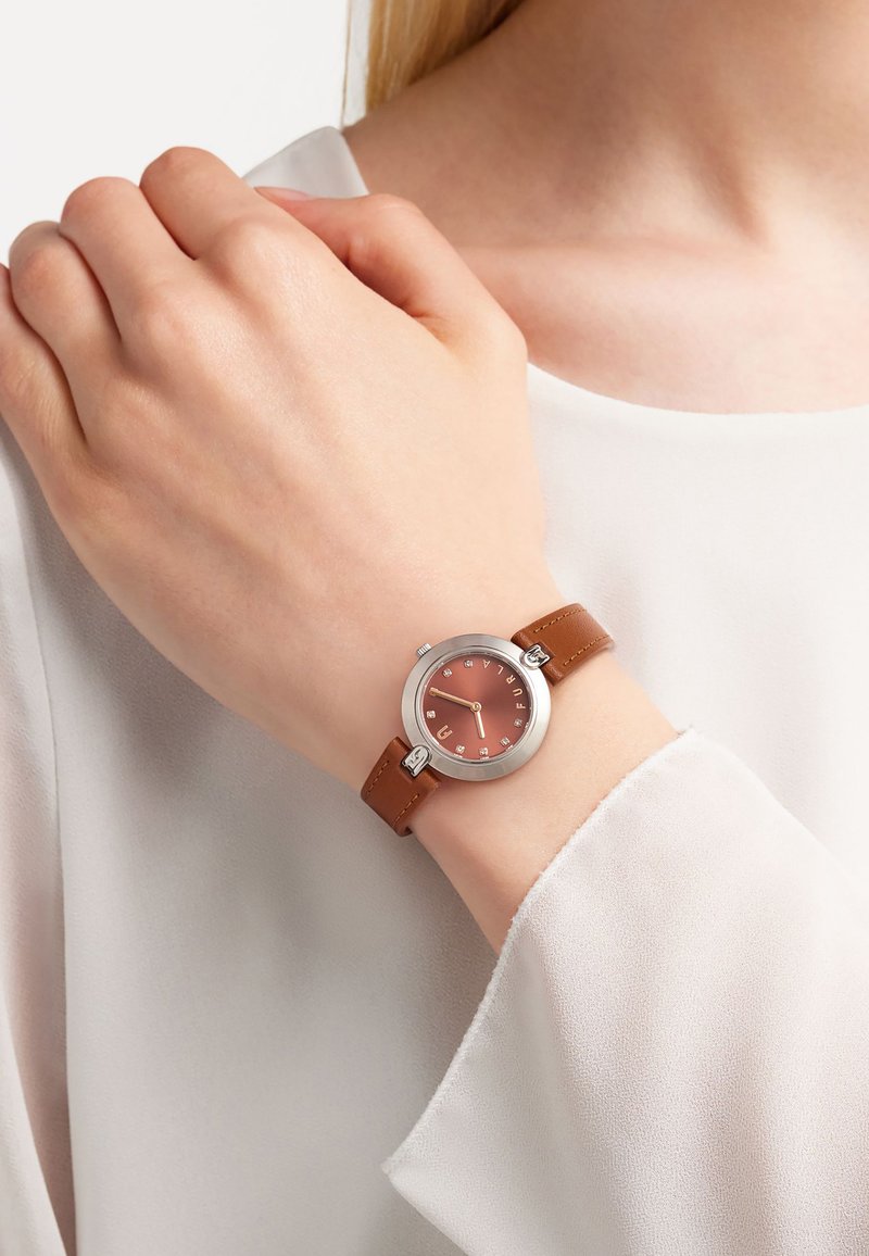 Montre à bracelet en cuir marron avec un cadran rond en or rose, des accents argentés et des marqueurs d'heure clairs. Portée sur une main contre une blouse blanche.