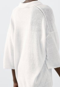 Weißer Pullover mit lockerem Schnitt, geripptem Kragen und strukturierter Strickoptik. Die Ärmel sind weit geschnitten und das Kleidungsstück verfügt über dezente Seitennähte.