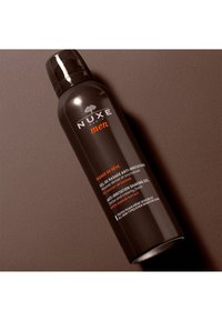 Svart cylindrisk flaska med en glänsande yta, märkt "NUXE men." Har vit och orange text som anger att det är en rakgel mot irriterad hud med baobabextrakt.