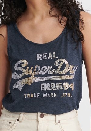 Kvinde iført en mørkeblå ærmeløs top med teksten "REAL SuperDry TRADE. MARK. JPN." og japanske tegn i hvid og guld.