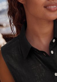 Schwarzes Kurzarmhemd mit Kragen, ausgestattet mit Knopfverschluss und Spitzendetails auf den Schultern. Glatte Textur mit dezentem Glanz.