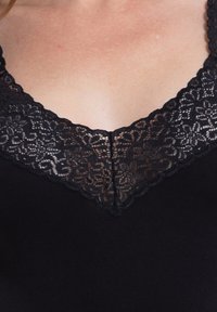 Schwarzes Kleidungsstück mit V-Ausschnitt, versehen mit zarter schwarzer Spitzenborte und gewellten Kanten, aus weichem, glattem Material.