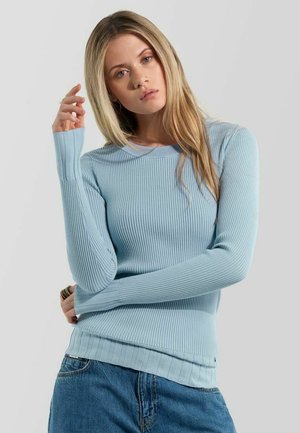 Junge Frau mit langen blondem Haar, die einen hellblauen, fein gerippten Pullover und blaue Jeans trägt, posiert mit einem verschränkten Arm und der Hand nahe am Gesicht.