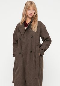 Brun houndstooth frakke med dobbeltbrystet design, store knapper og bælte detalje. Har en bred krave og en løs pasform.