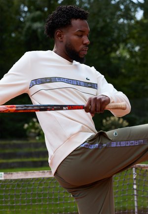 Bărbat îmbrăcat într-un hanorac alb și pantaloni oliv ținând o rachetă de tenis în fața unei fileu pe un teren de tenis în aer liber.