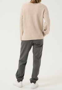 Helle beige Strickjacke mit entspannter Passform und überschnittenen Schultern, kombiniert mit dunkelgrauen schmal zulaufenden Hosen und weißen Turnschuhen.