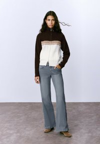 Chaqueta de punto con cremallera en un diseño de bloques de color marrón y crema, con textura de punto trenzado. Combinada con pantalones de mezclilla gris de pierna ancha.