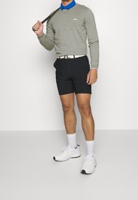 Pull en tricot gris clair, chemise bleue à col, short noir, ceinture blanche et chaussures de sport blanches avec des accents gris. Club de golf à la main.