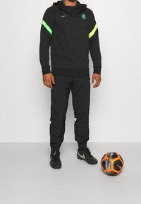 Haut de sport noir avec des accents vert fluo, pantalon noir et crampons. Un ballon de football noir et orange est au sol.