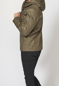 Mężczyzna poprawiający troczki oliwkowej kurtki z kapturem, ubrany w czarne spodnie, stojący na jednolitym jasnym tle.