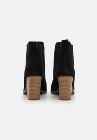 Botas negras de ante hasta el tobillo con punta redondeada, lengüetas para colocar, y un tacón de madera apilada. Superficie texturizada y sutiles detalles de costura.