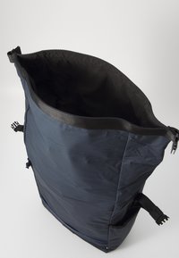 Mørkeblå ryggsekk med vid åpning, laget av glatt, vannavstøtende materiale, med sidestraps og en teksturert innvendig foring.