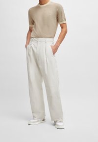Chemise en maille beige à manches courtes avec un col plus clair, associée à un pantalon large crème, présentant des plis devant et deux poches latérales.