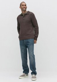 Brun ribbad stickad tröja med krage, tre knappar, kombinerad med blå denimjeans och ljusa sneakers. Modellen står mot en neutral bakgrund.