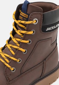 Bota de cuero marrón con cordones amarillos, ojales plateados y un cuello de tela negra. Presenta un diseño cosido y una suela de goma resistente.